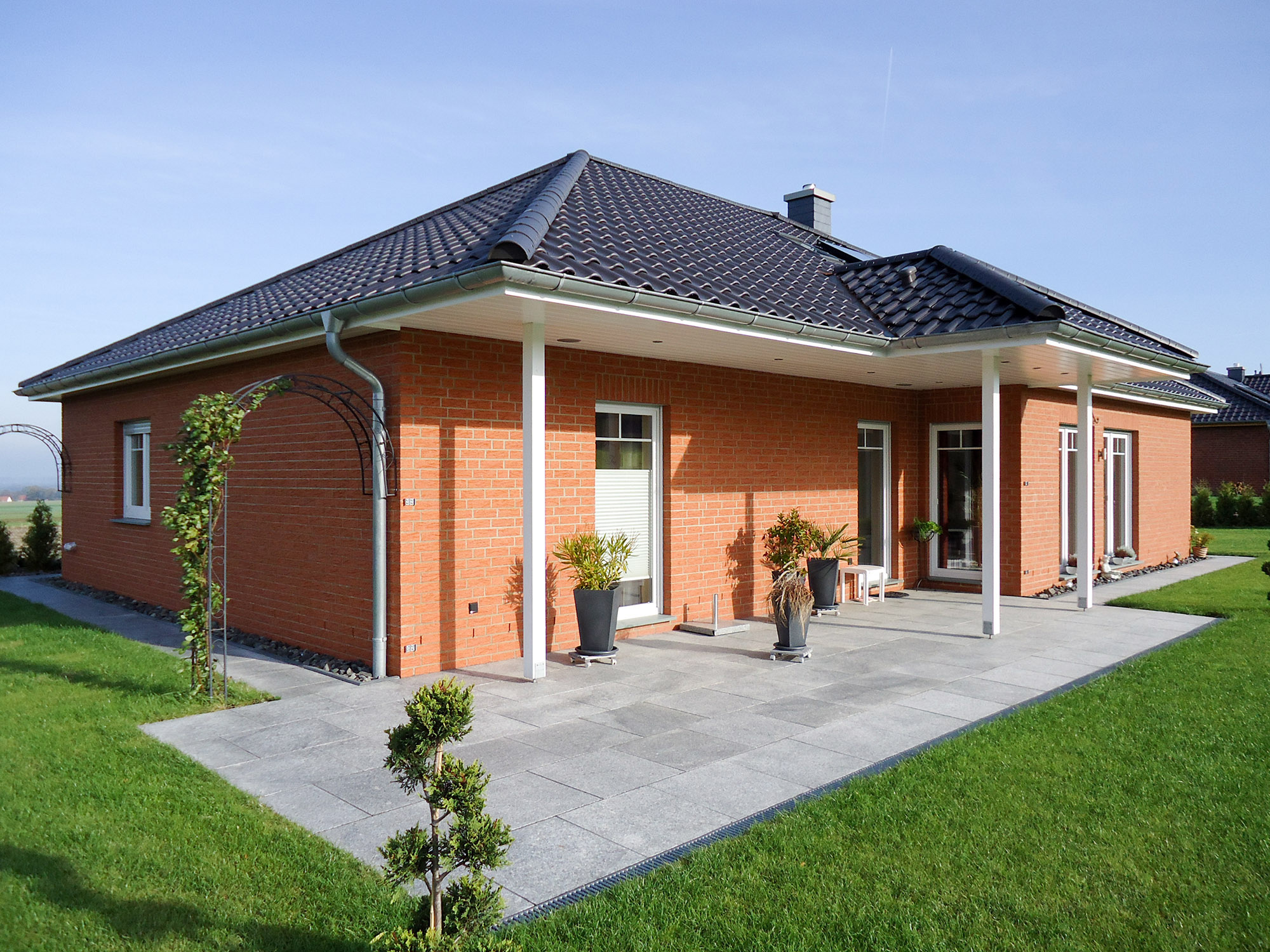 Moderner Klinkerbungalow mit überdachter Terrasse im Grünen Moderner Bungalow mit roter Klinkerfassade, Walmdach, überdachter Terrasse und gepflegtem Garten bei Sonnenschein.
