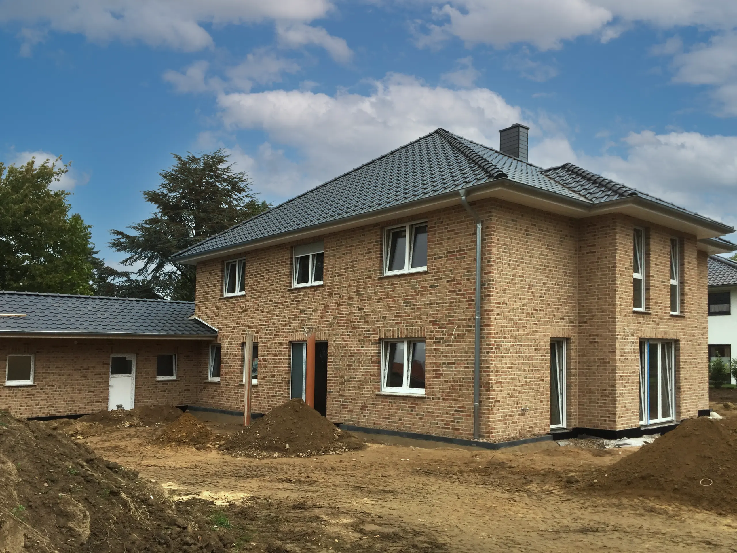 Klinkerhaus mit Walmdach, bodentiefen Fenstern und aufgeschütteter Erde im Vorgarten vor geplanter Gartengestaltung.