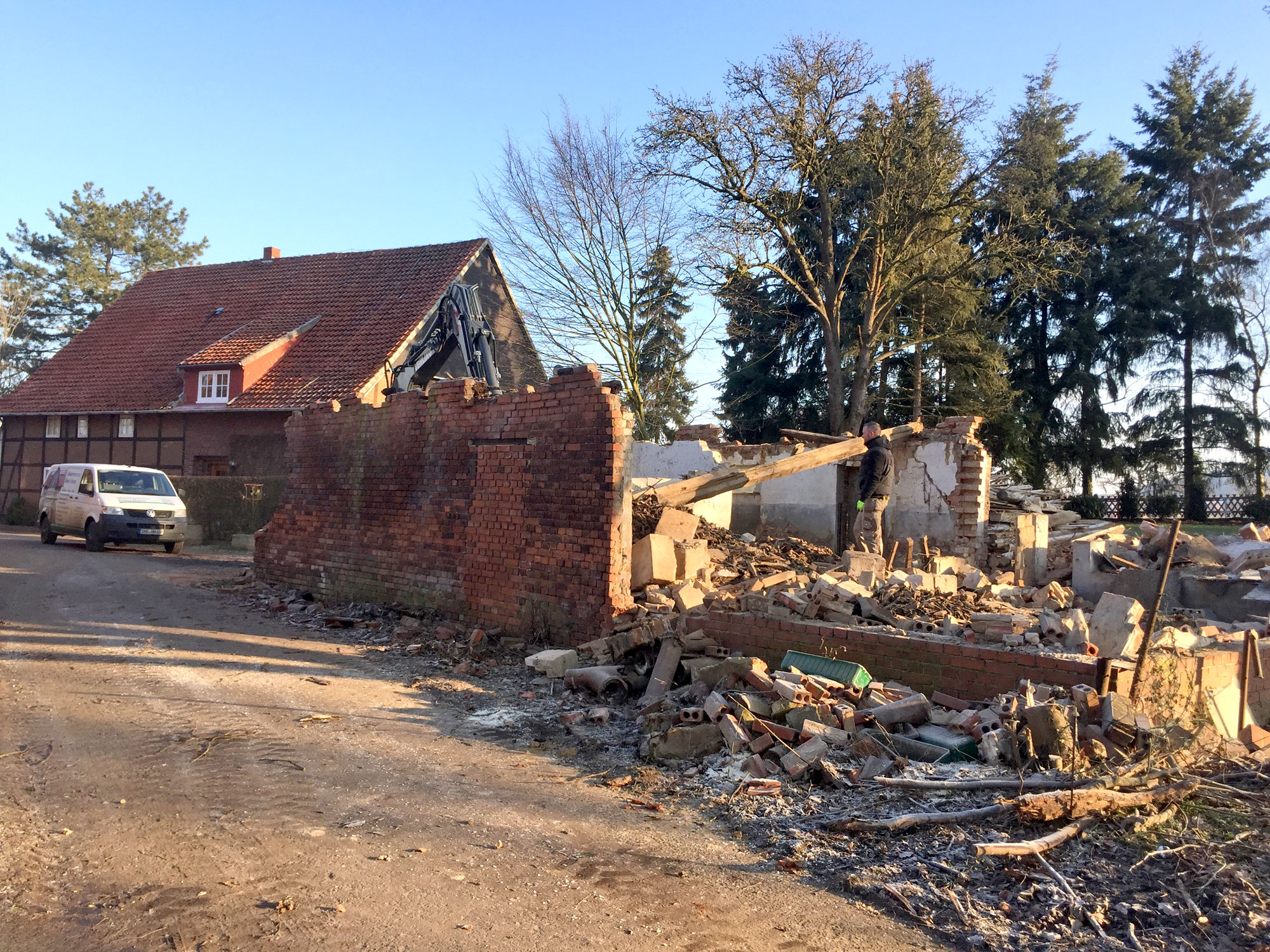 Teilabgerissenes Nebengebäude mit Bauschutt, Arbeiter trägt Holzbalken, im Hintergrund ein Fachwerkhaus und Bäume.