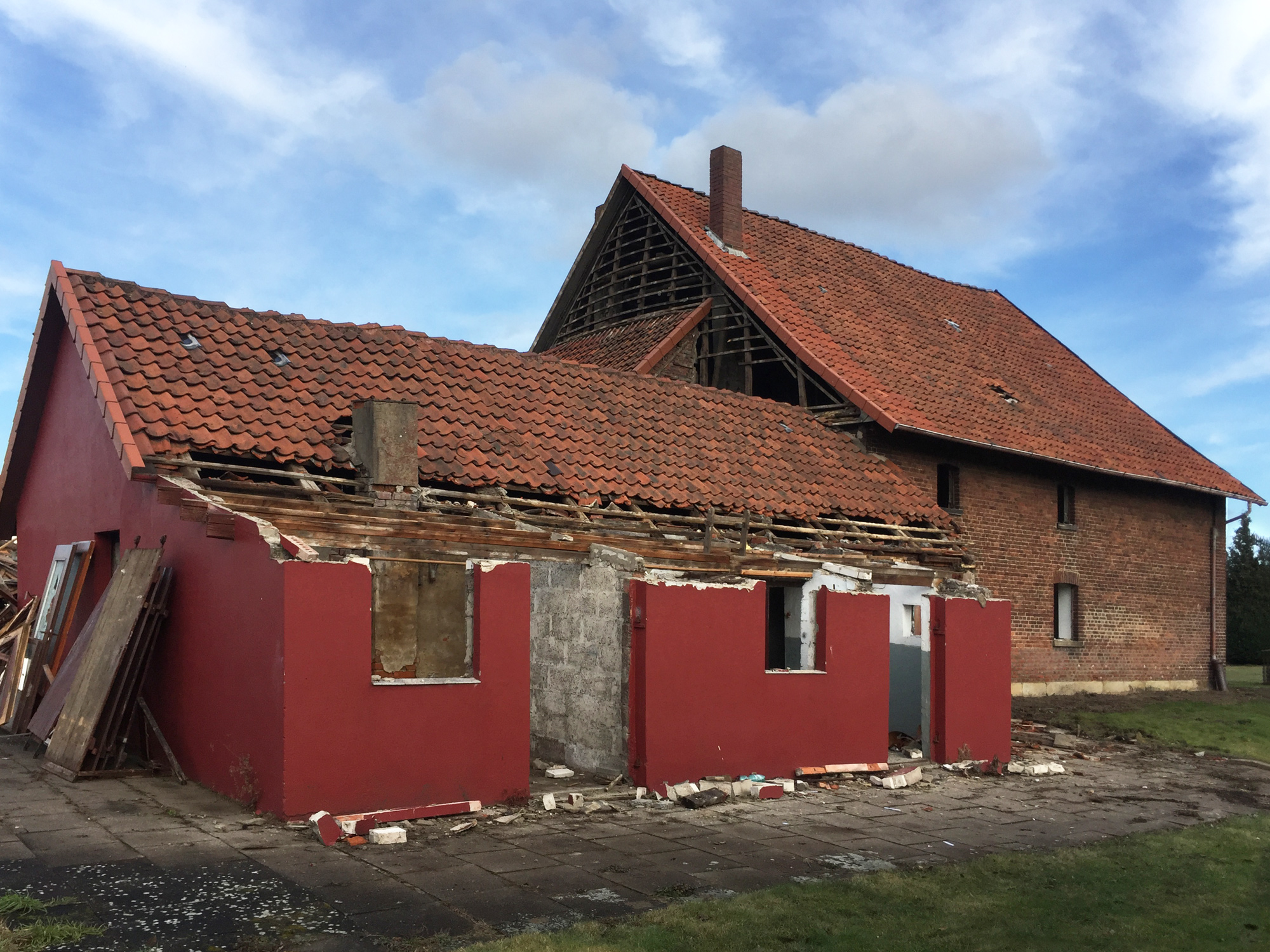 Teilweise abgerissenes Nebengebäude mit rotem Putz und abgedecktem Dach, dahinter ein größeres Wohnhaus aus Backstein.