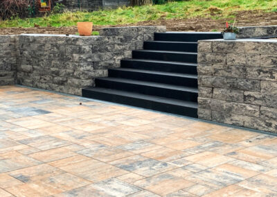 Gepflasterte Terrasse mit Natursteinmauer und schwarzer Treppe, die in einen höher gelegenen Garten führt.