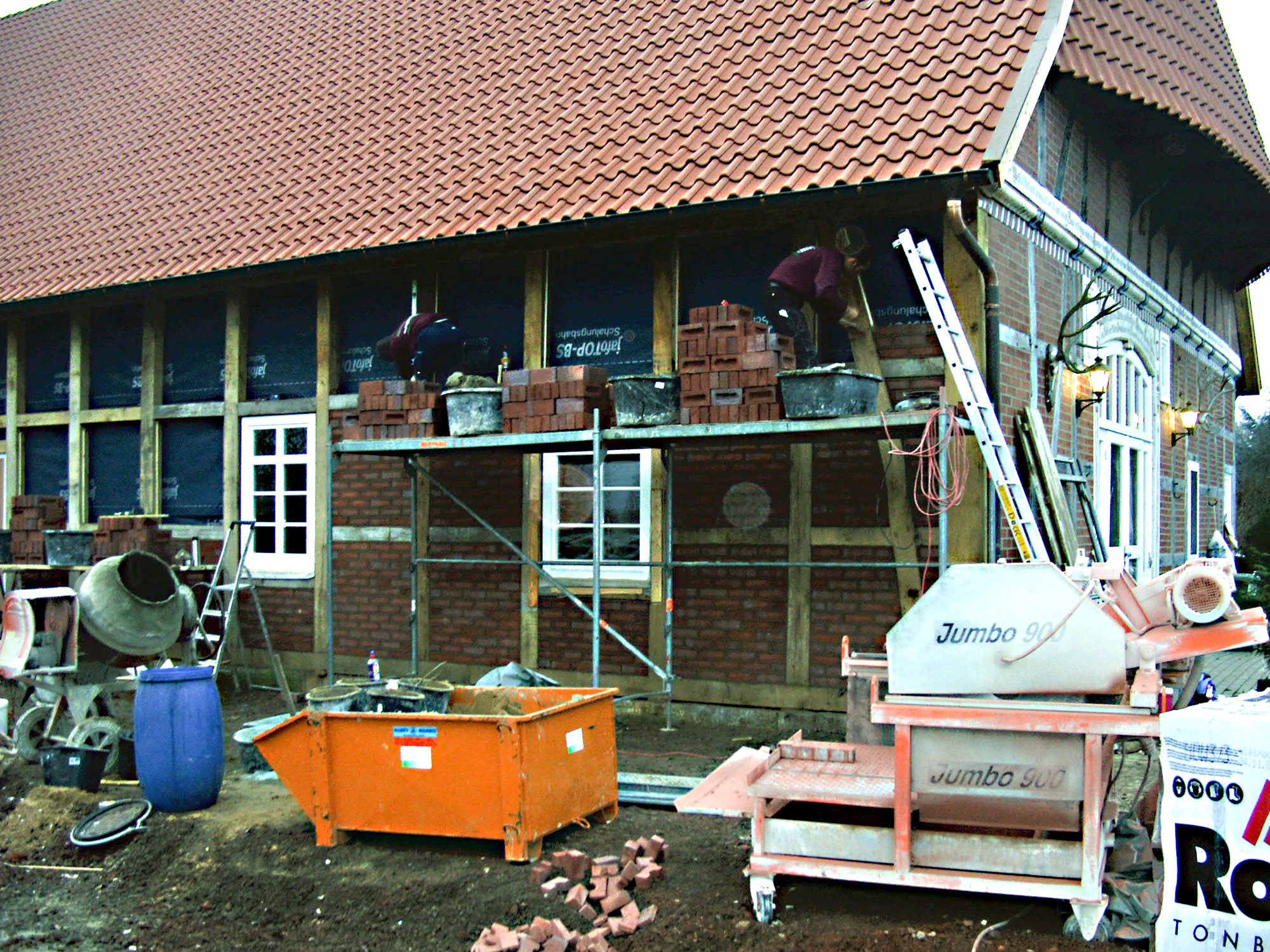 Handwerker arbeiten auf Gerüst an der Ausmauerung eines Fachwerkhauses mit Ziegeln, Baugeräten und Material im Vordergrund.