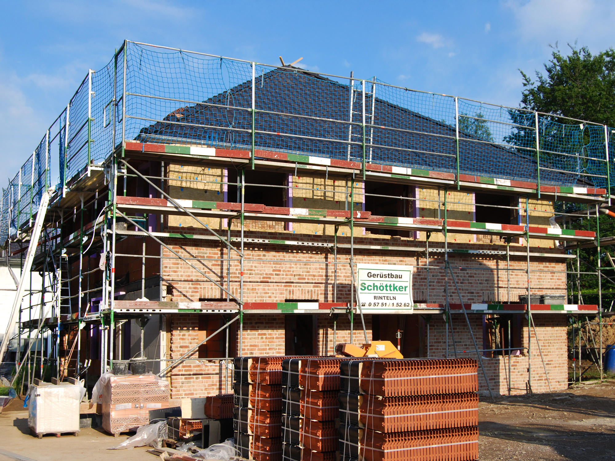 Mehrfamilienhaus im Rohbau mit Klinkerfassade, Gerüst und Dämmarbeiten; Baumaterialien liegen im Vordergrund bereit.