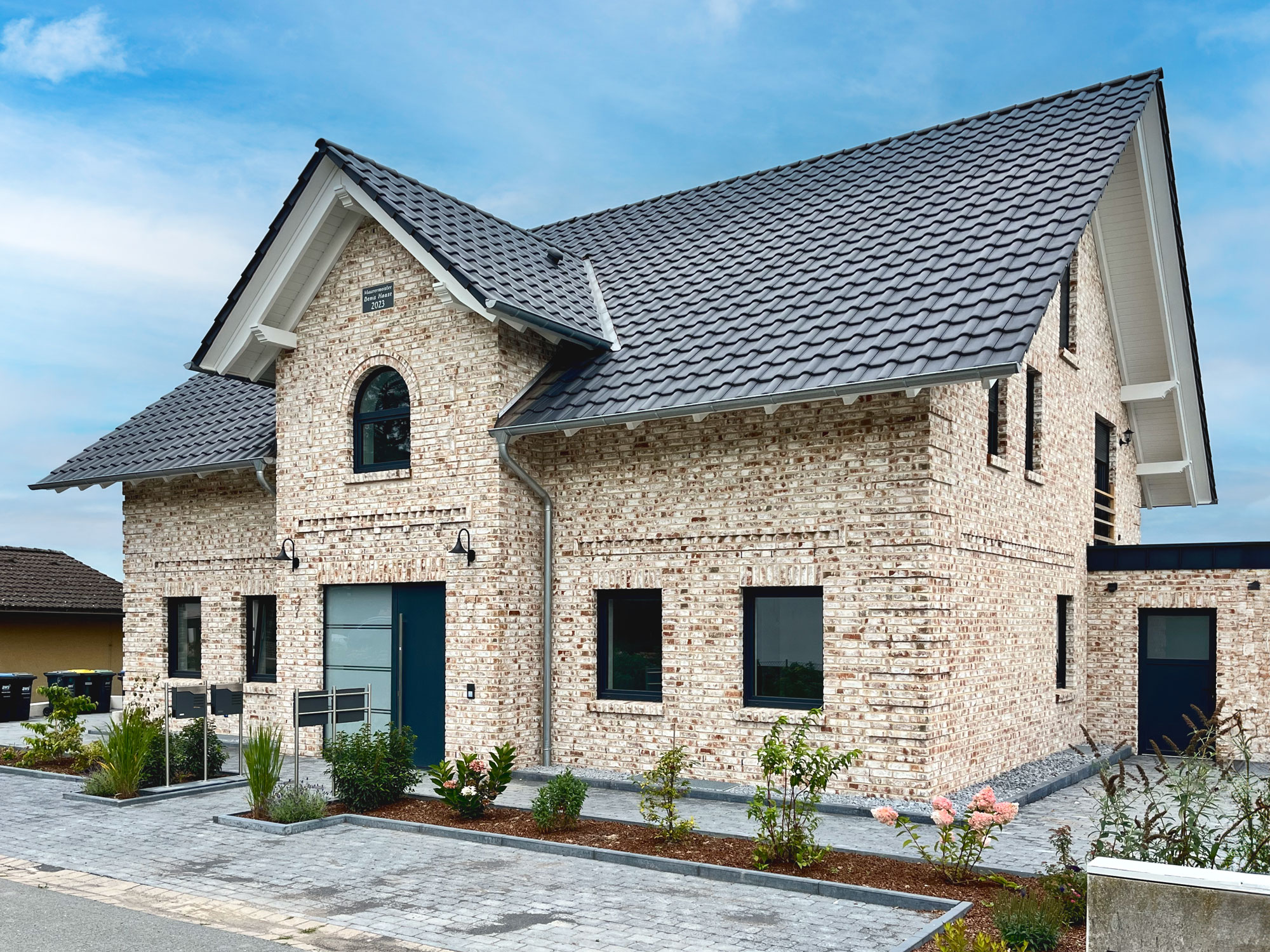 Einfamilienhaus mit heller Klinkerfassade und dunklem Ziegeldach in traditioneller Bauweise Zweigeschossiges Haus mit heller Klinkerfassade, dunklem Dach und gepflasterter Fläche mit Parkplätzen im Vordergrund.