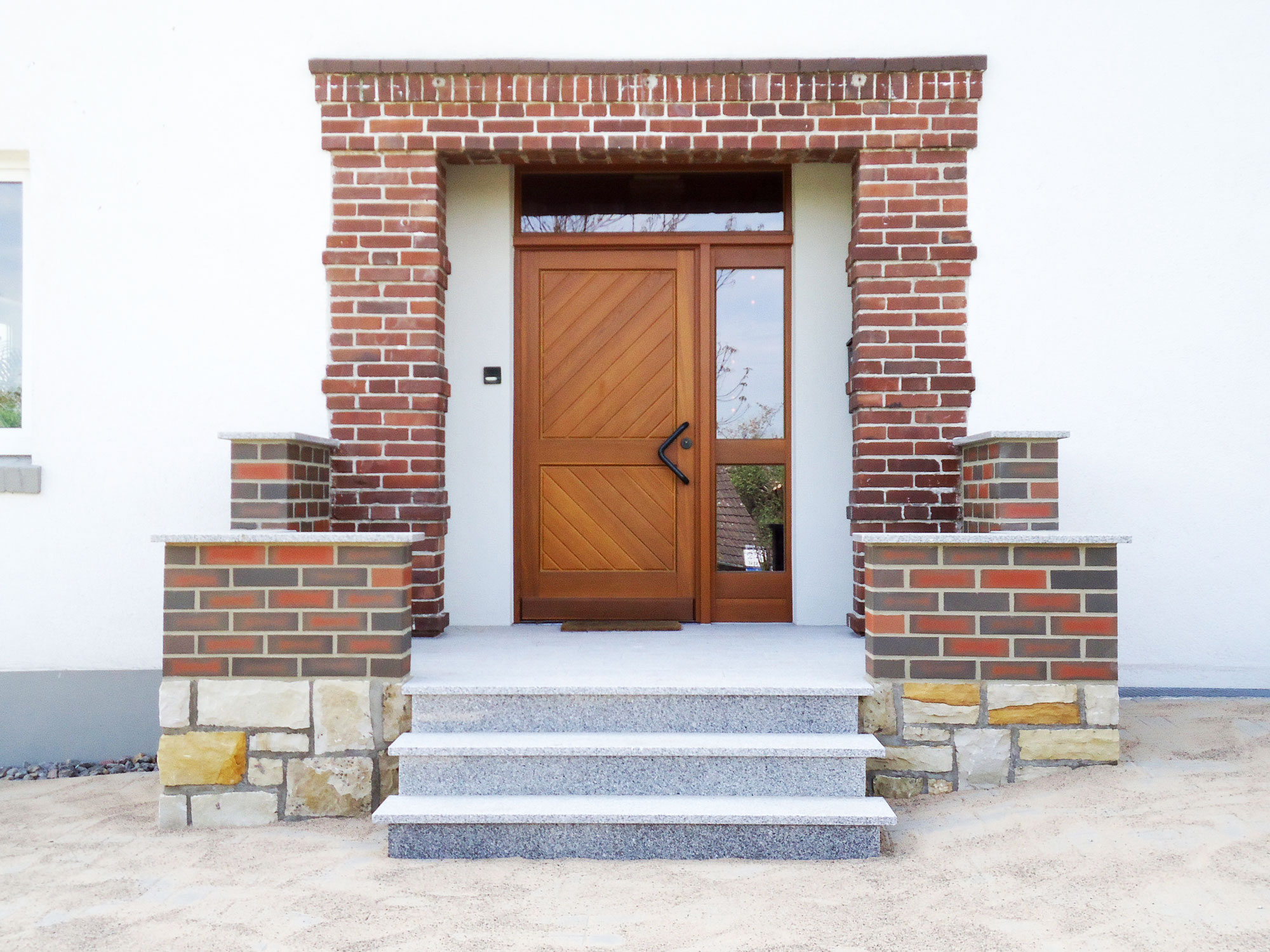 Sanierter Hauseingang mit Treppe und neuer Holzhaustür, rotem Klinkerrahmen und Natursteinmauerwerk auf heller Putzfassade