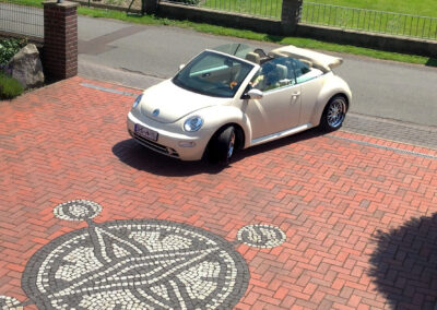 Hofeinfahrt mit rotem Pflasterstein und rundem Mosaik, auf den ein cremefarbener VW Beetle Cabrio bei Sonnenschein einfährt.