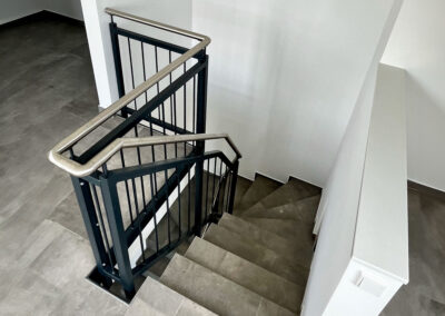 Moderne Innentreppe mit grauen Fliesen, mit anthrazitfarbenem Metallgeländer und Edelstahl-Handlauf in einem hellen Innenraum.