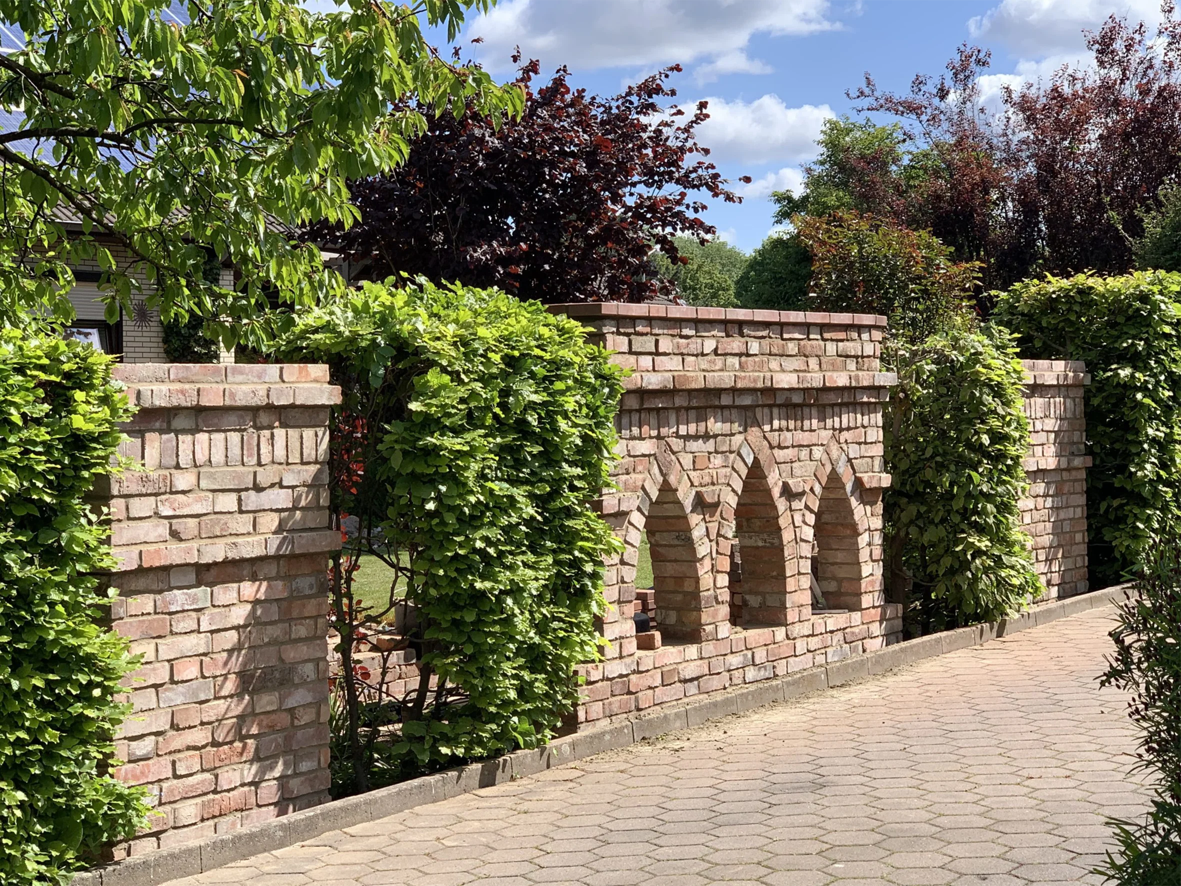 Gartenmauer aus Backstein mit gotischen Bögen und grüner Bepflanzung entlang eines gepflasterten Weges bei Sonnenschein