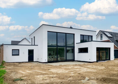 Modernes zweigeschossiges Einfamilienhaus im Bauhausstil mit Flachdach, großen Fensterfronten und weißer Fassade mit unbepflanzter Gartenanlage unter blauem Himmel mit kleinen Wolken. Links und rechts befinden sich Wohnhäuser.