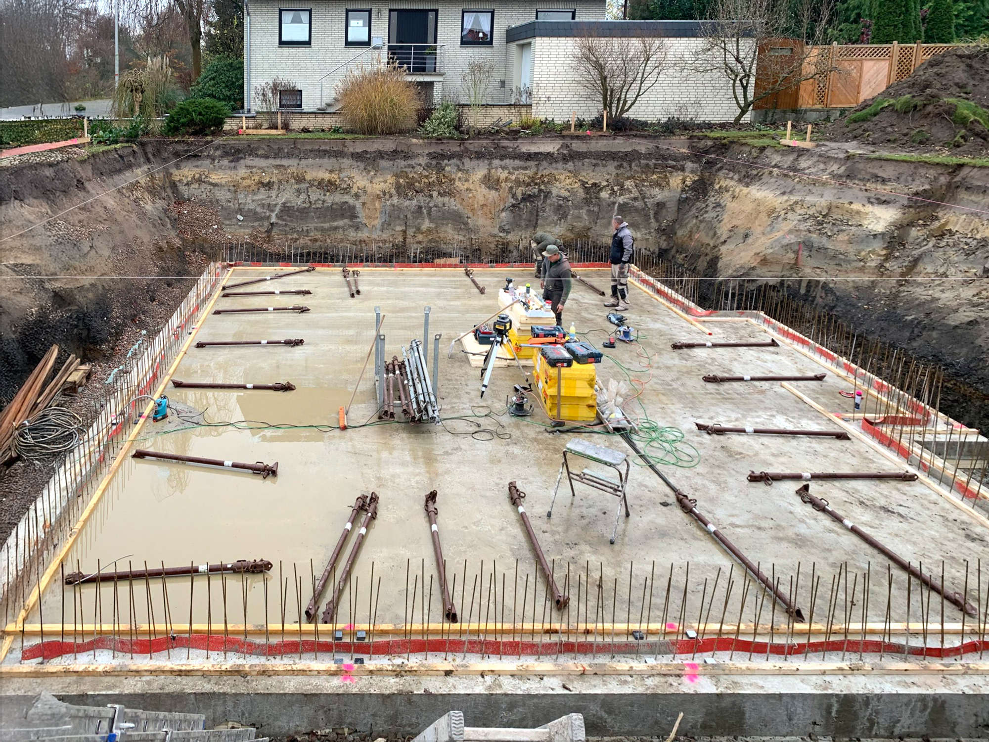 Verlegung einer Sohlplatte mit Bewehrung auf einer Baustelle im Wohngebiet Zwei Arbeiter auf Baustelle bei der Bewehrung einer Sohlplatte, Baugrube umgeben von Erde, Wohnhäuser im Hintergrund.