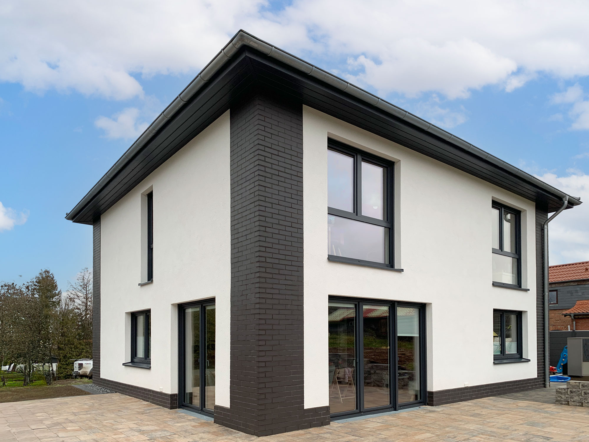 Moderne Stadtvilla mit weißer Putzfassade und dunklen Klinkerelementen Modernes zweigeschossiges Wohnhaus mit weißem Putz, dunklen Klinkerecken, dunkelgrauen Fenstern und gepflasterter Terrasse.