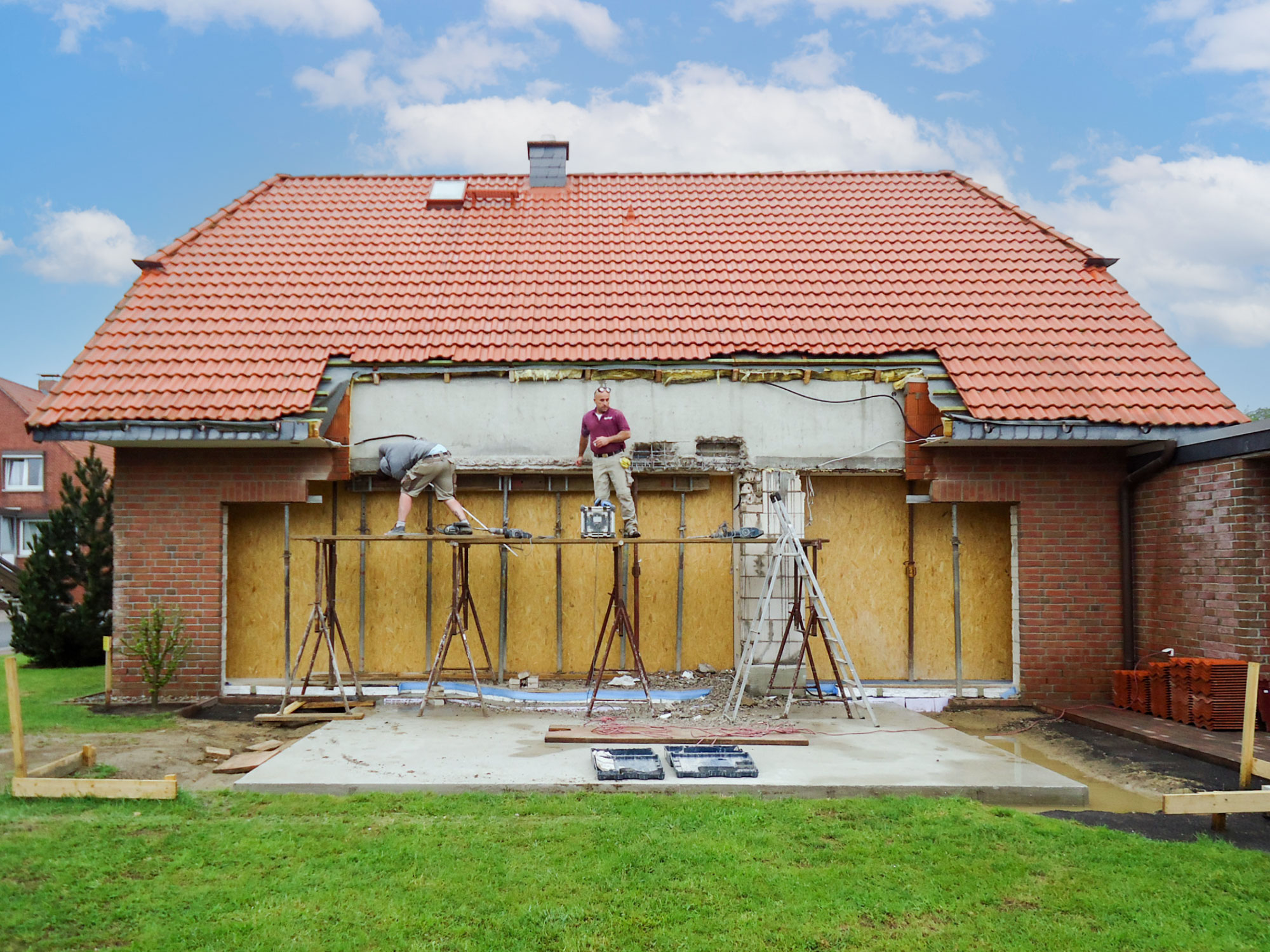 Haus mit rotem Dach, bei dem die Rückwand für einen Anbau geöffnet wurde, zwei Handwerker arbeiten am Rohbau.