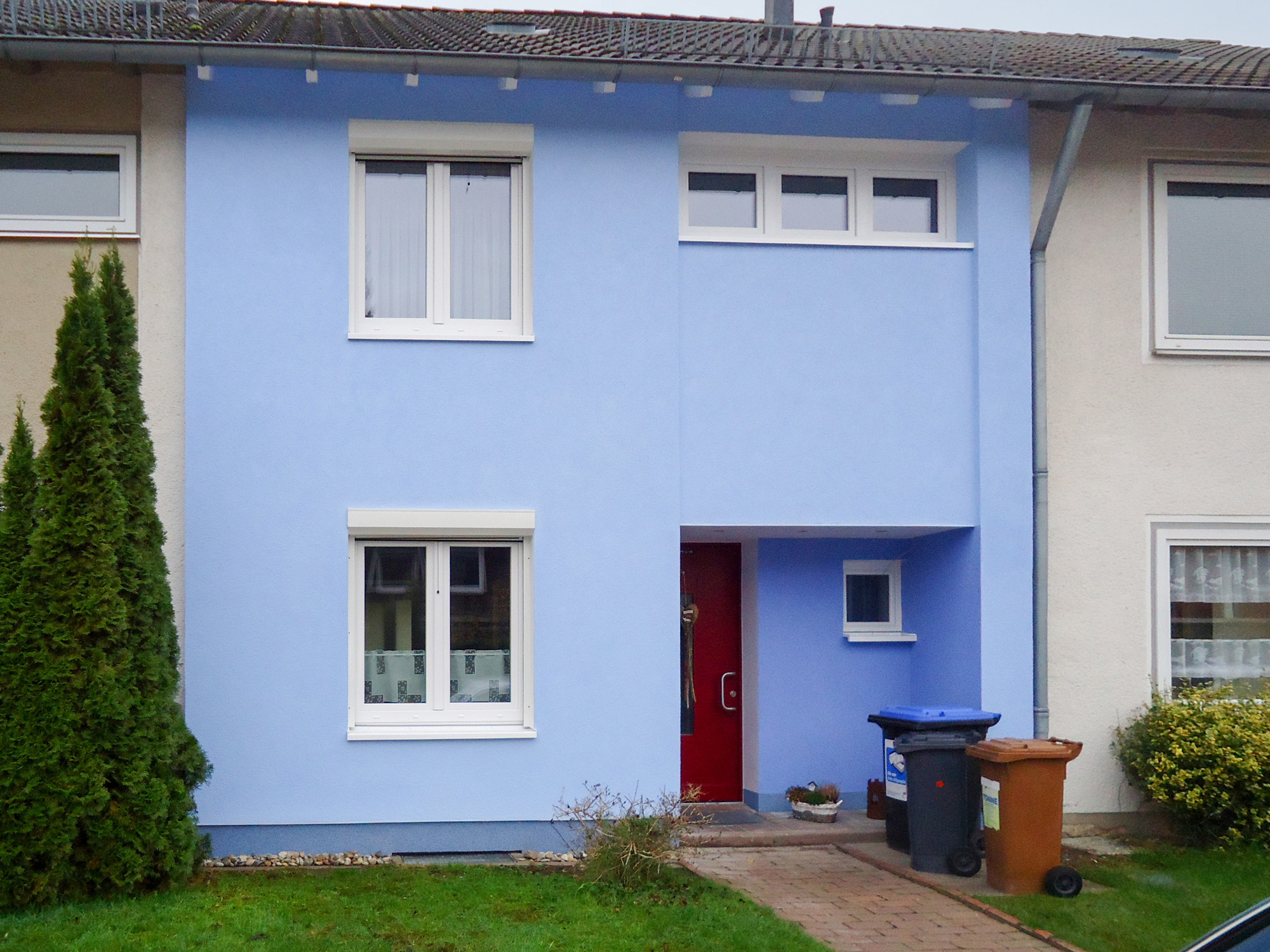 Frisch sanierte Reihenhausfassade mit hellblauem Putz, weißen Fenstern, roter Tür und Mülltonnen am Eingangsweg.